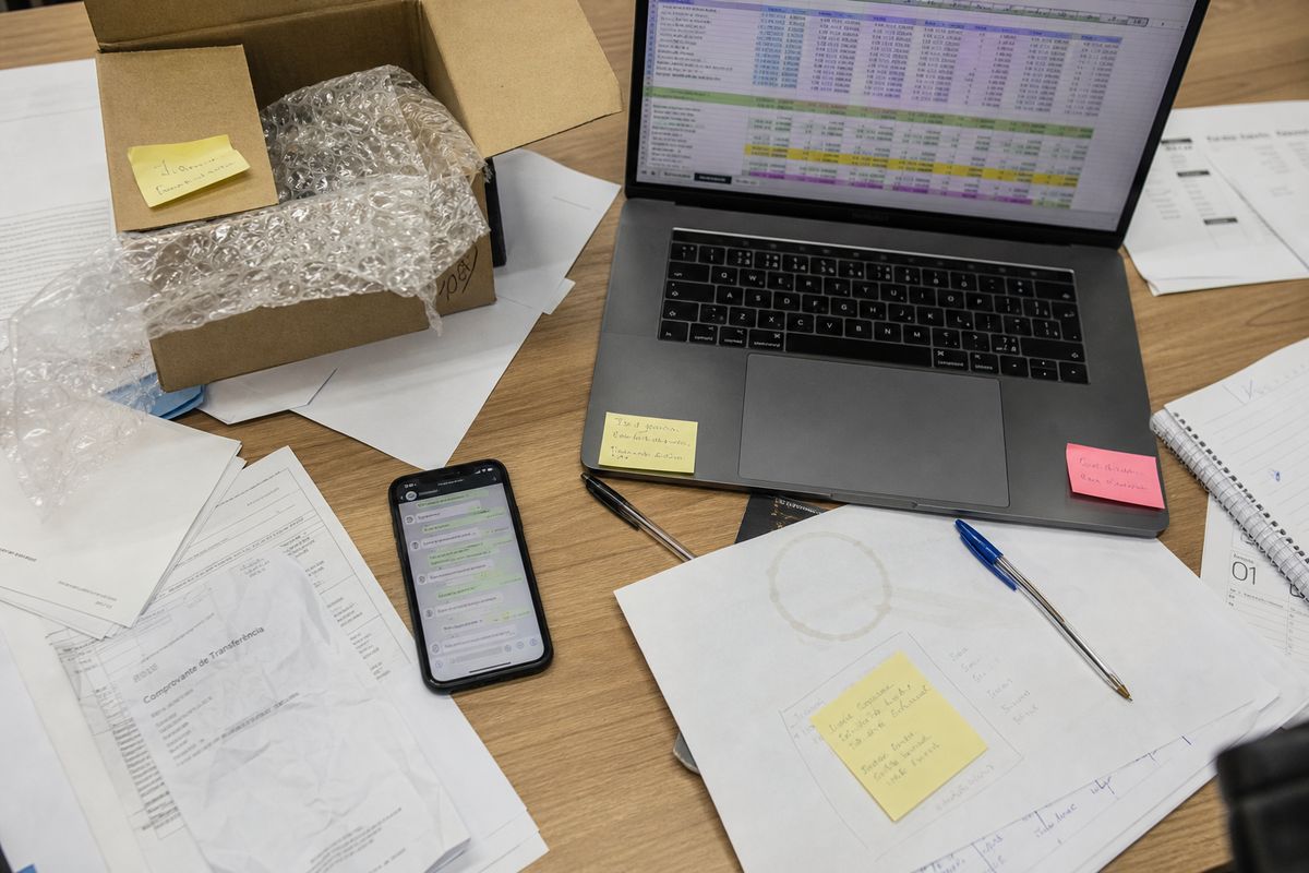 Mesa com caixa de amostra, planilha no laptop, papéis e celular espalhados